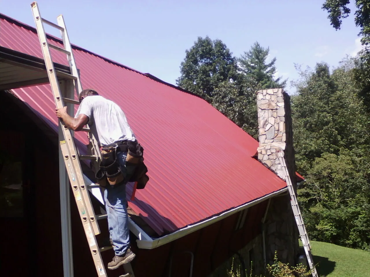 Roofer on a ladder beside a red metal roof and stone chimney — Soffit & Fascia Repair by Roofing Brothers in Wildomar